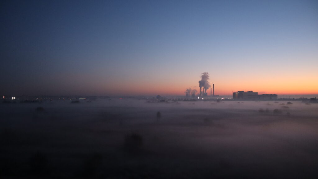 Low fog sits across a park, with a power station in the background. The sky is early morning blue.