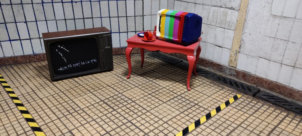 A TV set sits on the ground next to a red table with a portable TV on it, painted with the test card. 
