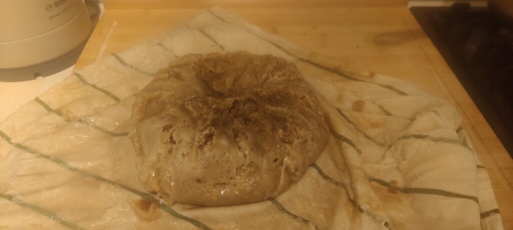 The dumpling sitting on the unwrapped 'clootie.' The pudding looks pale, and is covered in a wet skin caused by the flour.
