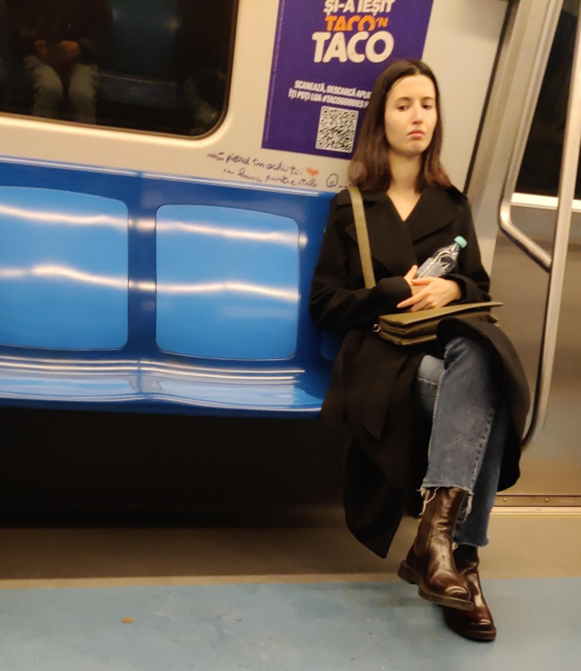A slightly sad looking young woman sits on a seat on the Bucharest metro, looking downwards. Contemplative look. Has a handbag on her lap and a bottle of water in her right hand.